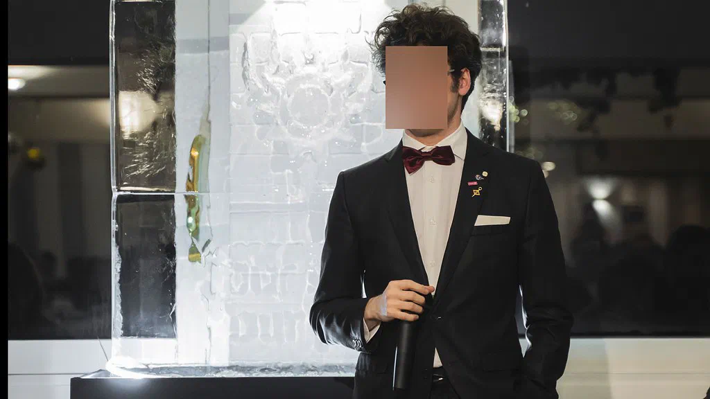 The same photograph after the anonymization pass: the man's face is entirely hidden under a flat, featureless patch, while the suit, bow tie, microphone and ice sculpture are untouched.