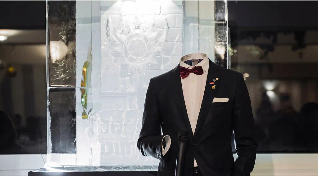 A dark suit with a white shirt, deep red bow tie and pocket square stands at a microphone in front of a carved, backlit ice sculpture. There is no person in the clothes: the collar opens onto empty interior space, the cuff is hollow, and the garments hold their shape as if still worn.