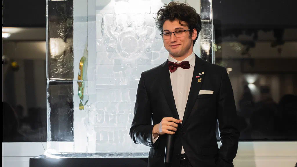 A man in a dark suit with a deep red bow tie, glasses and curly hair holds a microphone in front of a backlit ice sculpture, smiling slightly.